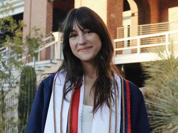 Woman with dark brown hair stands outside a brick building with graduation gown and cords