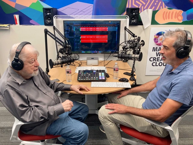 A man in a gray shirt and jeans, wearing headphones, sits across from another man in blue shirt and khakis in a podcast recording studio