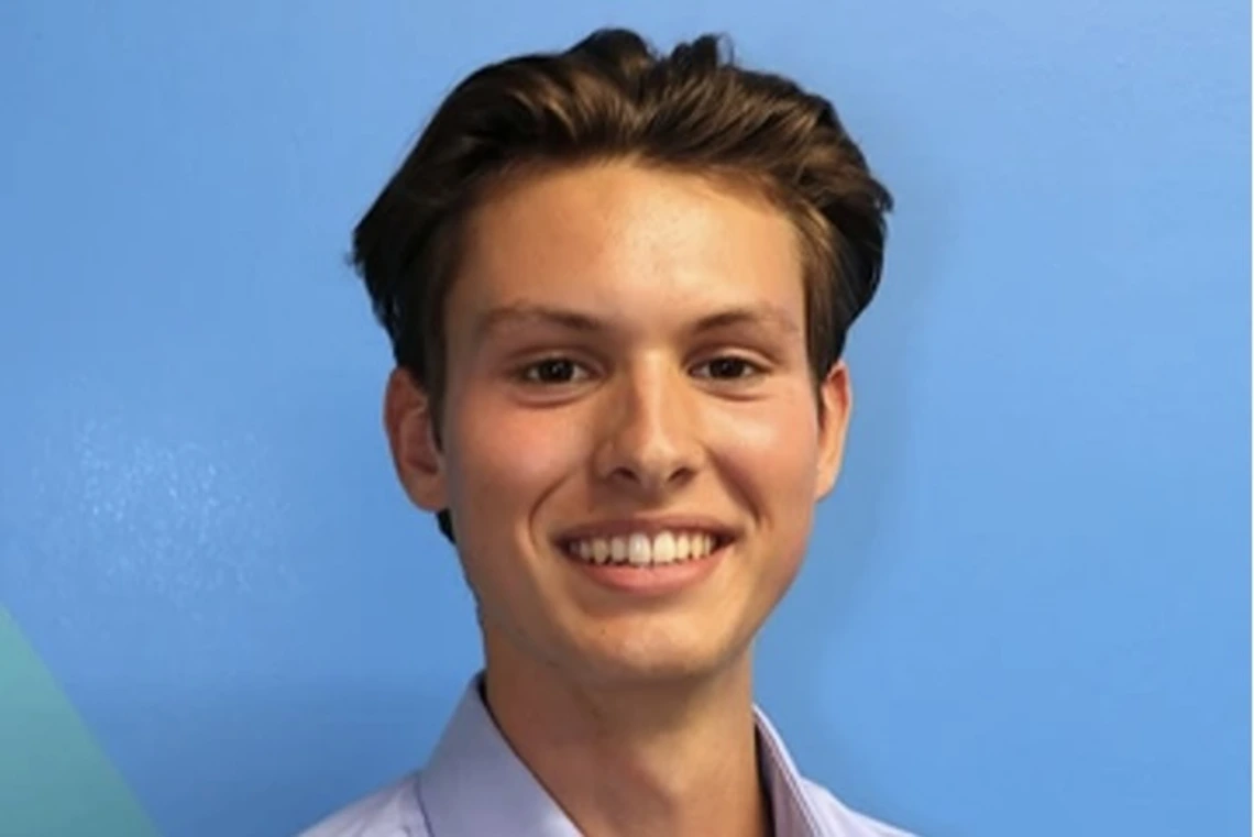 Young man with dark blonde hair and wearing a blue collared shirt smiles against a blue background