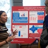 Female student with long brown hair, stands in front of a posterboard about food insecurity, gesturing while talking to a community member